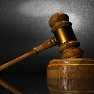 May include: A wooden gavel with a gold-colored striking head rests on a matching wooden block. The gavel handle extends to the left, and the background is a dark, textured surface with a spotlight.