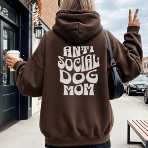 Könnte beinhalten: Ein brauner Kapuzenpullover mit dem Text "ANTI SOCIAL DOG MOM" in weißer, stilisierter Schrift. Der Kapuzenpullover hat eine Kapuze und lange Ärmel. Die Person trägt einen schwarzen Rock und hält eine Kaffeetasse.