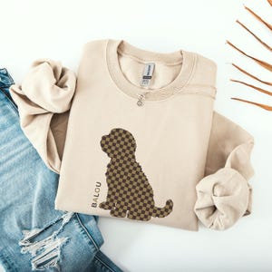 Puede incluir: Sudadera beige con una silueta de perro a cuadros marrón y beige. La palabra "BALOU" está impresa verticalmente en el lado izquierdo del perro. Un collar plateado con un pequeño colgante está en la sudadera. La sudadera está al lado de unos vaqueros azules.
