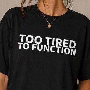 Puede incluir: Camiseta negra con el texto blanco "TOO TIRED TO FUNCTION". La persona lleva vaqueros azules y un collar dorado. Un estampado enmarcado de Matisse cuelga de la pared detrás de la persona.