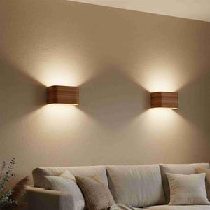 May include: Two wooden wall sconces emitting a warm glow. The rectangular sconces have a curved top and bottom, mounted on a neutral wall. The light shines downwards and outwards, creating a cosy ambiance. The sconces are above a sofa, creating a warm atmosphere.
