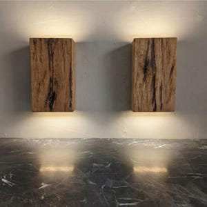 May include: Two wooden wall sconces with a natural wood grain finish. Each sconce is rectangular and emits a warm, downward-facing light. The sconces are mounted on a light gray wall above a dark, marbled surface.
