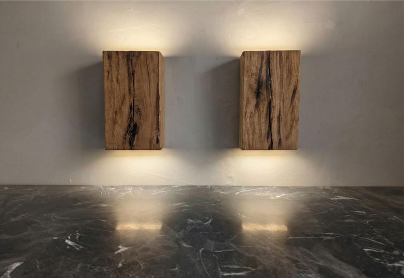 May include: Two wooden wall sconces with a natural wood grain finish. Each sconce is rectangular and emits a warm, downward-facing light. The sconces are mounted on a light gray wall above a dark, marbled surface.