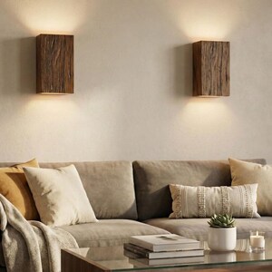 May include: Two rectangular wooden wall sconces with warm light illuminating the space. The sconces are mounted on a neutral-colored wall above a sofa with pillows and a blanket. A coffee table with books and a plant is in front of the sofa.
