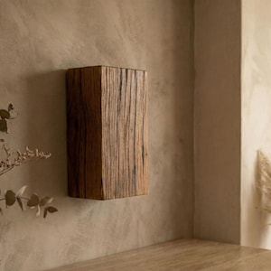 Op de afbeelding: Een rechthoekige houten wandplank met een natuurlijke, verweerde uitstraling. De plank is gemaakt van donkerbruin hout en is bevestigd aan een beige muur. De houtnerf is zichtbaar en voegt textuur toe. Het is een decoratief element.