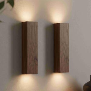 May include: Two rectangular wooden wall sconces in a warm brown finish. Each sconce emits a soft, downward-facing light, creating a cosy ambiance. The sconces are mounted on a light-coloured wall, showcasing their minimalist design.