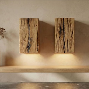 May include: Two rectangular wooden wall sconces with a natural, weathered appearance. The sconces are mounted on a light-colored wall and emit a warm, inviting glow from the bottom. The wood grain and texture are prominent.