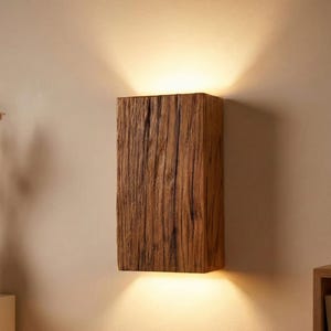 Puede incluir: Aplique de pared rectangular de madera con un acabado natural texturizado. El aplique emite luz hacia arriba y hacia abajo, creando un brillo cálido. La veta de la madera es visible, lo que añade encanto rústico. Montado en una pared de color neutro.