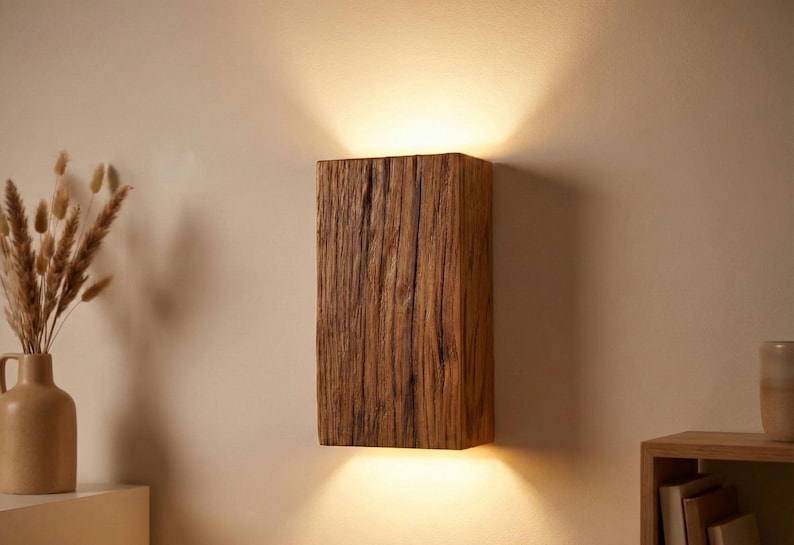 May include: A rectangular wooden wall sconce with a natural, textured finish. The sconce emits light upwards and downwards, creating a warm glow. The wood grain is visible, adding to its rustic charm. The sconce is mounted on a neutral-colored wall.