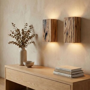 May include: Two wooden wall sconces with a natural wood grain texture are mounted on a beige wall. A wooden console table holds a vase with dried foliage, a small bowl, and a stack of books. The scene is illuminated by warm light.