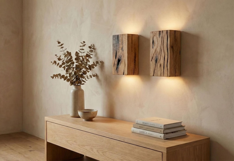 May include: Two wooden wall sconces with a natural wood grain texture are mounted on a beige wall. A wooden console table holds a vase with dried foliage, a small bowl, and a stack of books. The scene is illuminated by warm light.