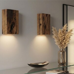 May include: Two rustic wooden wall sconces with warm lighting. The sconces are rectangular and made from textured wood. A glass shelf holds a decorative bowl and a vase with dried plants. A mirror is in the background.