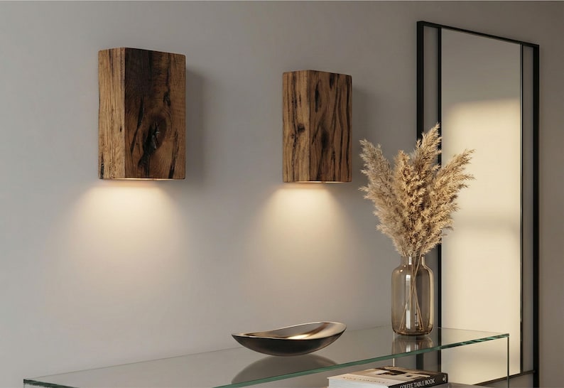 May include: Two rustic wooden wall sconces with warm lighting. The sconces are rectangular and made from textured wood. A glass shelf holds a decorative bowl and a vase with dried plants. A mirror is in the background.