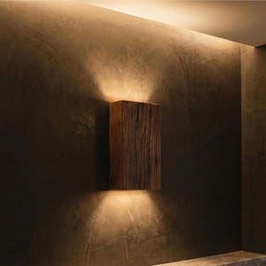 May include: A rectangular wooden wall sconce with a natural, textured finish. The sconce emits warm light upwards and downwards, creating a cozy ambiance. The fixture is mounted on a dark, textured wall.