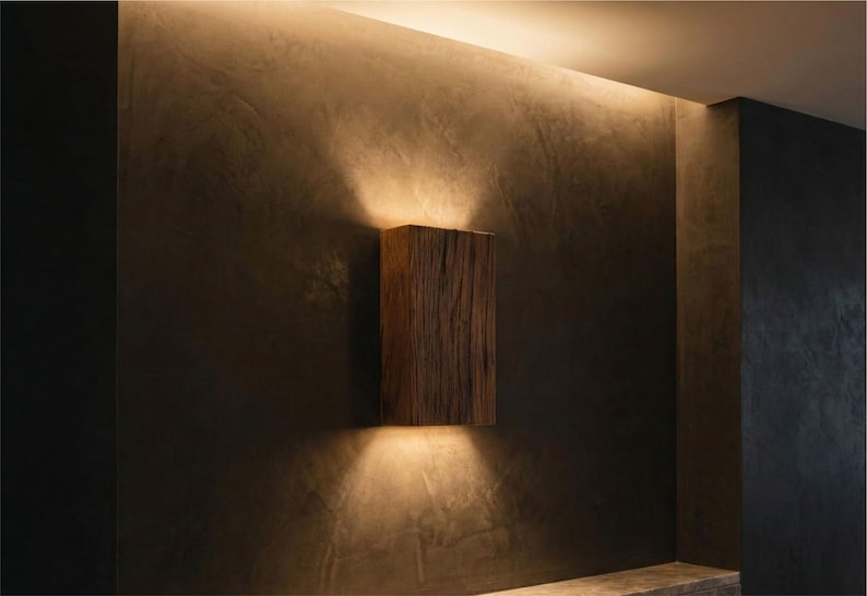 May include: A rectangular wooden wall sconce with a natural, textured finish. The sconce emits warm light upwards and downwards, creating a cozy ambiance. The fixture is mounted on a dark, textured wall.