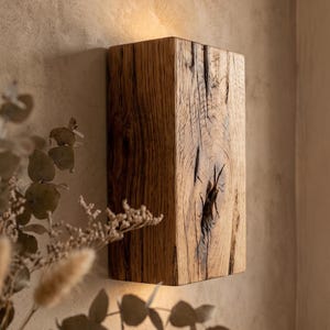 May include: A rectangular wall sconce crafted from a solid block of natural wood with a prominent dark knot in the center. The fixture emits a warm, soft glow from both the top and bottom edges, highlighting the textured grain of the timber.