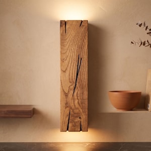 May include: A rectangular wooden wall sconce with a warm, natural wood grain finish. The sconce has a vertical design with a crack-like detail and emits a soft, downward glow. The sconce is mounted on a beige wall.