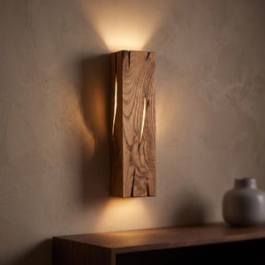 May include: A rectangular wall sconce crafted from natural wood with a prominent grain pattern. The fixture features vertical fissures that allow warm light to emit from the top, bottom, and sides, casting a soft glow against the wall surface.