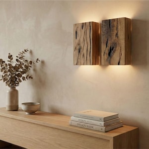 May include: Two wooden wall sconces with a warm glow, mounted on a neutral-toned wall. The sconces are made of textured wood, showcasing natural grain patterns. A wooden console table below holds a vase, bowl, and books.