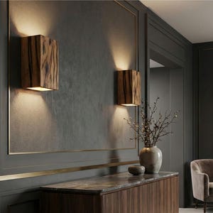 May include: Two wooden wall sconces with a warm, natural wood grain pattern are mounted on a dark gray wall. The sconces emit a soft, downward glow, creating a cozy ambiance. The room features a dark wood cabinet and a neutral-toned armchair.