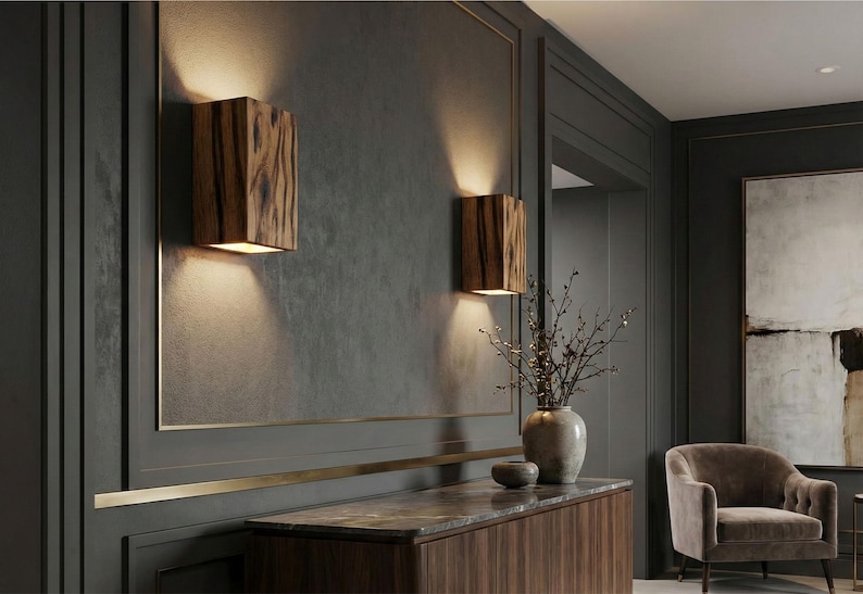 May include: Two wooden wall sconces with a warm, natural wood grain pattern are mounted on a dark gray wall. The sconces emit a soft, downward glow, creating a cozy ambiance. The room features a dark wood cabinet and a neutral-toned armchair.