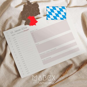 Könnte beinhalten: Ein weißes Planerblatt mit dem Datum "01. Januar 2026" und Zeitschlitzen, mit einer Deutschlandkarte und der bayerischen Flagge im Hintergrund. Der Planer liegt auf einem beigen Stoff, mit Weizenähren. Der Markenname "MABEX homecore" steht unten.