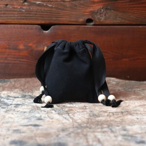 Könnte beinhalten: Ein kleiner, schwarzer Kordelzugbeutel mit abgerundeter Form. Die Tasche hat schwarze Bänder und ist mit weißen Holzperlen und schwarzen Quasten verziert. Die Tasche steht auf einer strukturierten Oberfläche mit Holzgrund.