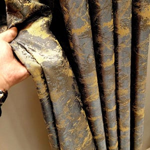 Könnte beinhalten: Nahaufnahme eines luxuriösen Vorhangstoffs mit strukturiertem Muster. Der Stoff weist eine Mischung aus Gold- und Grautönen auf, die ein reichhaltiges und elegantes Aussehen erzeugt. Der Vorhang wird offen gehalten und zeigt das detaillierte Gewebe und Design.