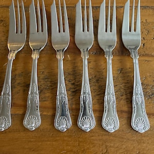 May include: Six silver forks with detailed, ornate handles. The forks are arranged in a row, displaying their tines and decorative features. The handles have a shell-like design, adding a touch of elegance to the silverware.