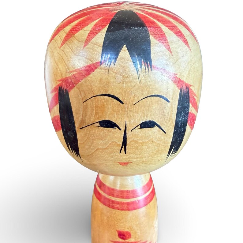 Peut inclure: Une poup&eacute;e kokeshi en bois avec une grande t&ecirc;te arrondie, des cheveux noirs peints &agrave; la main, des sourcils arqu&eacute;s et des yeux &eacute;troits. Le grain du bois naturel est visible, rehauss&eacute; par des motifs floraux rouges et des bandes horizontales.