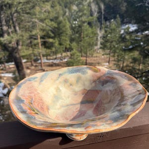 May include: A handmade ceramic bowl with a unique, organic shape. The bowl features a blend of colors, including beige, blue, and red, with a glossy finish. The rim has a wavy design, and the bowl is supported by a small, rounded base.