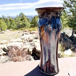 May include: A tall ceramic vase with a flared rim, featuring a glossy glaze that blends deep chocolate brown, dark blue, and creamy white tones in a vertical, flowing drip pattern that extends from the top down to the base of the vessel.