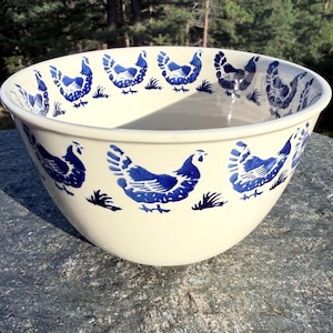 May include: A large, round, cream-colored ceramic mixing bowl featuring a repeating pattern of blue hens or chickens around the exterior and interior rim. The bowl has a smooth, glossy finish and a wide, open top with a simple, rounded edge design.