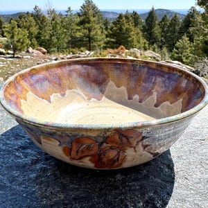 May include: A large, handmade ceramic bowl with a wide rim. The bowl features a blend of colors, including tan, brown, and purple, with a leaf design on the side. The bowl is sitting on a dark gray stone surface.