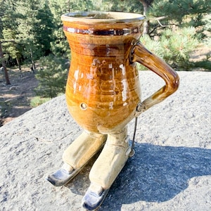 May include: A ceramic mug with a glossy brown glaze, featuring a rounded body and a single handle. The mug is supported by two sculpted ceramic legs wearing light-colored ski boots attached to dark-colored skis, creating a standing figure.