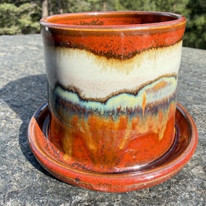 May include: A cylindrical ceramic planter with a matching saucer, featuring a multi-colored glaze with horizontal bands of deep orange, creamy white, and a soft teal blue, showing a glossy finish with visible drips and blended color transitions.