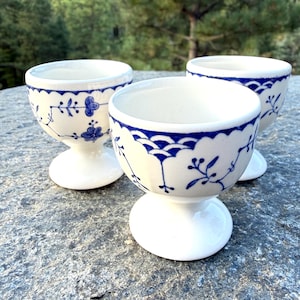 May include: Three white ceramic egg cups featuring a blue hand-painted floral and scalloped pattern. Each cup has a rounded bowl shape supported by a sturdy pedestal base. The design includes delicate blue vines and small flower motifs.