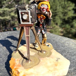 May include: A figurine of a clown with yellow hair, a black top hat, and a black coat with star-patterned lapels, standing behind a vintage bellows camera on a wooden tripod. The clown holds a small orange object near their face.