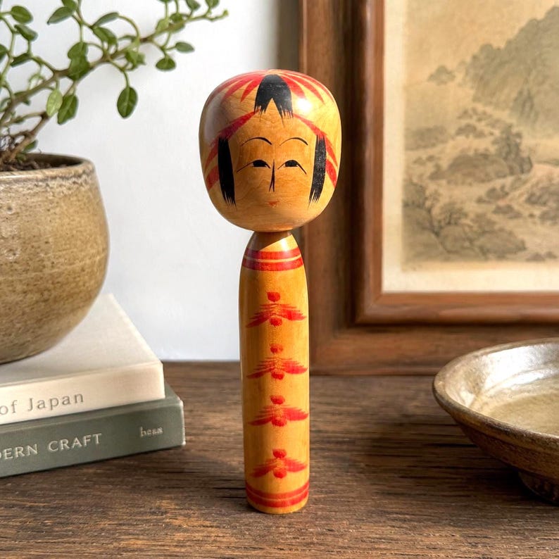Peut inclure: Une poup&eacute;e Kokeshi en bois avec une t&ecirc;te arrondie et un corps cylindrique. La t&ecirc;te pr&eacute;sente des cheveux noirs peints et des accents rouges, tandis que le corps est orn&eacute; d'une s&eacute;rie verticale de motifs floraux rouges et de bandes rouges.