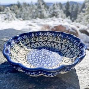 May include: A round, scalloped-edge ceramic bowl with a blue and white floral pattern. The bowl has a dark blue rim and a detailed design on the interior. The background shows a snowy landscape.