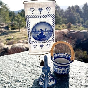 May include: A decorative display featuring a blue and white porcelain bell, a small bucket with a woven handle, and a framed porcelain plate with a nautical scene. The frame is white with blue cross-stitch hearts.