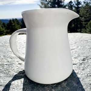 May include: A white ceramic pitcher with a curved handle and a spout. The pitcher is sitting on a gray stone surface with a natural background of trees and a blue sky. The pitcher has a smooth, glossy finish.