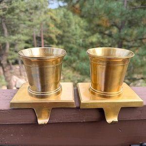 May include: Two polished brass stocking holders, each featuring a cylindrical cup-shaped top mounted on a flat square base with a curved hook extending downward from the front edge to secure the item onto a mantel or shelf surface.