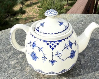 Pode incluir: Um bule de cerâmica branca com um padrão floral azul pintado à mão com vinhas e flores delicadas. O bule inclui uma tampa a condizer com um puxador, um bico curvo e uma pega lateral robusta, tudo decorado com detalhes em azul.