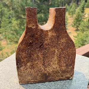 May include: A U-shaped ceramic vase with a textured, mottled brown and tan finish. The object features a flat base, wide sides that taper slightly toward the top, and a deep, rounded central cutout between two vertical pillars at the opening.