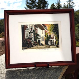 May include: A framed print of a street scene with buildings and trees. The print is in a dark wood frame with a white mat. The print depicts a street with buildings, trees, and flowers. The text "Dawn Mermaid Street, Peggy" is visible.