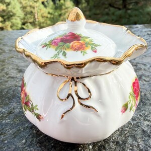May include: A white porcelain sugar bowl with a matching lid, featuring a scalloped rim with gold trim. The body and lid are decorated with colorful red and yellow rose floral patterns. A gold-painted bow detail adorns the front of the bowl.