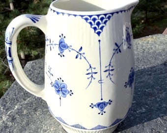May include: A white ceramic pitcher featuring a blue floral pattern with delicate vines and leaves. The rim and handle are decorated with blue scalloped details and lines. The body has a ribbed texture and a curved handle on the side.