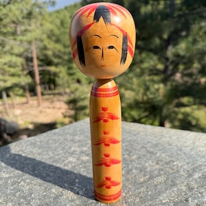 Peut inclure: Une poup&eacute;e Kokeshi en bois avec une t&ecirc;te arrondie, des cheveux noirs peints &agrave; la main, des accents rouges et un visage simple. Le corps cylindrique est orn&eacute; d'un motif floral rouge r&eacute;p&eacute;titif et de bandes horizontales sur un bois clair.