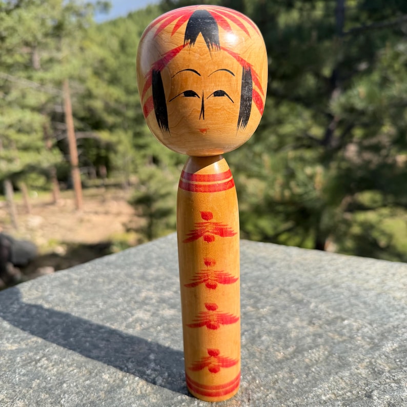 Peut inclure: Une poup&eacute;e Kokeshi en bois avec une t&ecirc;te arrondie, des cheveux noirs peints &agrave; la main, des accents rouges et un visage simple. Le corps cylindrique est orn&eacute; d'un motif floral rouge r&eacute;p&eacute;titif et de bandes horizontales sur un bois clair.